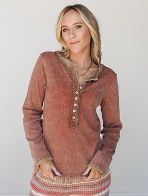 A lived-in mineral-washed henley top in warm chocolate brown, styled with a striped skirt and layered gold necklaces — the kind of top that looks like it's been loved for years.