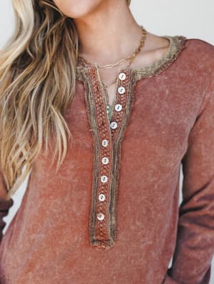 A close-up of a rust-toned mineral-washed henley top with layered crochet trim and pearl buttons down the placket — the kind of top that looks like it has a history, styled with delicate gold layered necklaces.
