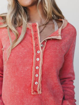 A close-up of a washed ribbed henley top in a warm carrot red, styled open at the collar with a layered gold chain necklace — the kind of top that looks like it's already your favorite.