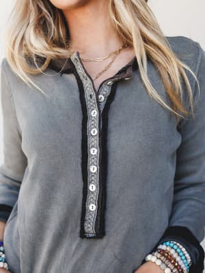 A close-up of a washed slate-grey ribbed henley top with a crochet-trimmed button placket and contrast black edges — the kind of effortless layering piece that looks just as good with jeans as it does under a flannel.