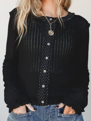 A black mixed-texture cardigan with a polka dot button-front panel and ruffle trim details, styled casually over light wash jeans with layered gold necklaces — the kind of top that looks like you tried just enough.