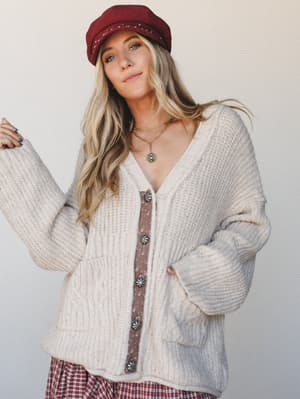 A cream chunky knit cardigan with a contrast button placard and concho-style buttons, styled with a plaid skirt and deep red baker boy cap — the kind of effortless fall layering that looks like it took no thought but absolutely did.