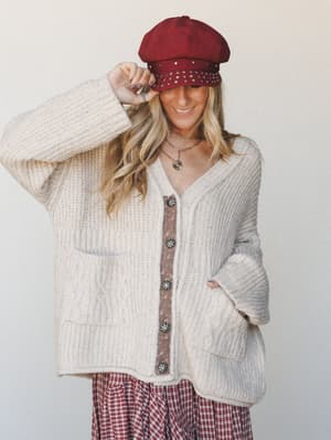 A smiling model tips the brim of a studded burgundy newsboy cap while layered in a chunky cream knit cardigan over a floral cami and plaid midi skirt — the whole look has that effortless fall-afternoon energy.