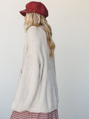 Shot from behind, a model in an oversized cream knit cardigan and red plaid skirt gazes softly over her shoulder — the kind of cozy-collected look that feels like the first cool day of fall finally arrived.