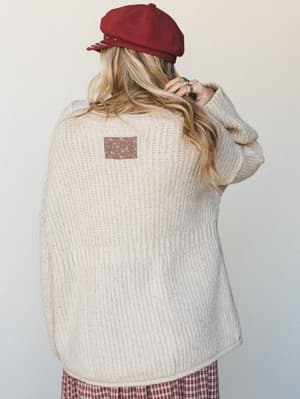 Shot from behind, a model in an oversized oatmeal knit cardigan with a back patch detail pairs it with a crimson baker boy cap and plaid skirt — the kind of effortless layering that feels like a Saturday morning with nowhere to be.