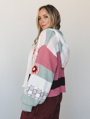 A model shot from the back-turned-three-quarter angle shows a richly textured patchwork cardigan in dusty pinks, sage green, white, and deep burgundy — the kind of sweater that looks like it has a whole story to tell, finished with crochet flower app