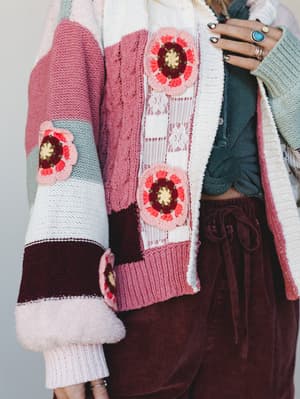 A close-up of a patchwork knit cardigan layered over a tied teal top and burgundy velvet pants — the crochet flower appliqués and mixed knit textures make it feel like something you'd find at a vintage market and never let go of.