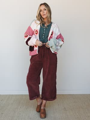 A relaxed full-body shot of a model layering a chunky color-block crochet floral cardigan over a teal henley top, paired with wide-leg corduroy pants in deep burgundy and brown mules — the kind of effortless fall outfit that looks like it came togeth