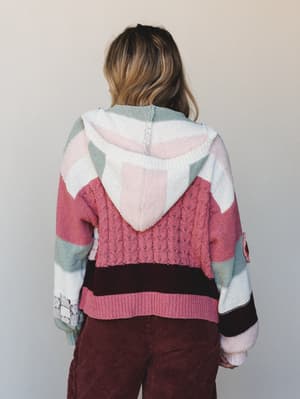 A back-view shot of a mixed-knit hooded cardigan in a patchwork of rosy pinks, sage green, cream, and deep burgundy — the kind of sweater that looks like it has a whole personality and a good story to go with it, paired with rust-toned corduroy pants