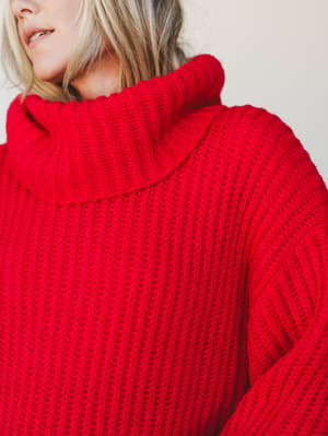 A close-up of an oversized ruby red turtleneck sweater dress, showing the chunky ribbed knit texture and draped cowl neck in rich detail — cozy confidence in its purest form.