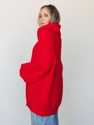 A cozy, oversized ruby red turtleneck sweater dress shot in profile against a soft white studio wall — the kind of piece that makes staying in feel like a whole look.