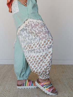 A close-up side view of mixed-media harem-style pants layered with a sheer floral lace overlay panel, paired with a green striped top and rainbow crochet buckle sandals — the kind of outfit that looks like it was assembled with a lot of personality a