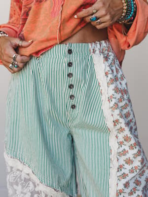 A close-up of mixed-print wide-leg pants layered with a cropped coral top, heavy silver and turquoise ring stacks, and beaded bracelets — the kind of outfit that looks like it was assembled with intention and a lot of personality.