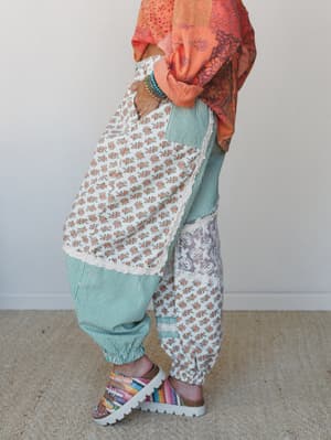 A side-profile shot of patchwork harem-style pants mixing teal stripes, ditsy florals, and a muted botanical block print — styled with an orange printed top, stacked beaded bracelets, and colorful flatform sandals for a wonderfully collected boho loo