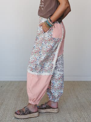 A side-profile shot of patchwork harem-style pants mixing ditsy floral and orange stripe panels with lace trim, styled with a graphic tee, stacked bead bracelets, and leopard print platform sandals — the kind of pants that make people ask where you g