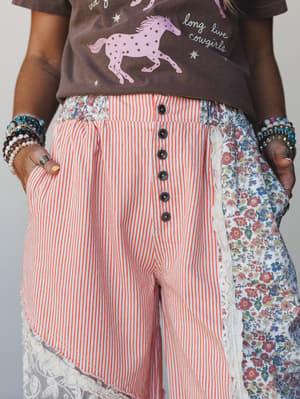 A close-up of mixed-media wide-leg pants combining orange pinstripe fabric, floral side panels, and lace trim at the hem — styled with a graphic cowgirl tee and stacked beaded bracelets for a playful, eclectic boho look.
