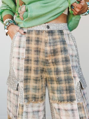 A close-up detail shot of mixed-fabric cargo pants in faded camel plaid paired with a mint green cropped sweatshirt, styled with layered turquoise and pearl bracelets for a worn-in, expressive boho look.