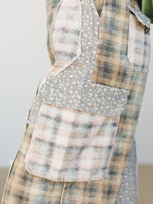 A close-up detail shot of wide-leg patchwork pants mixing a soft camel and blush plaid with a grey ditsy floral fabric panel, showing a patch pocket and raw-edge seam detailing — the kind of piece that looks like it was made from a grandmother's quil