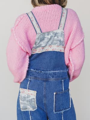 Person wearing blue denim overalls with floral patterns and a pink sweater on a plain background