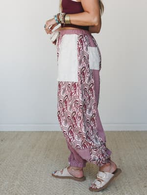A side-profile model shot of relaxed patchwork joggers in mauve and cream paisley — stacked with turquoise and gold bracelets, paired with a burgundy crop top and white buckle sandals, the whole look feels like it wandered in from somewhere good.