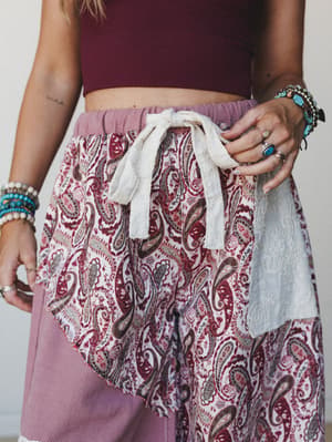 A close-up mid-shot of wide-leg joggers in a mauve and paisley patchwork fabric, styled with a cropped burgundy top and layered boho jewelry — the tie waist and embroidered panel details do all the talking.