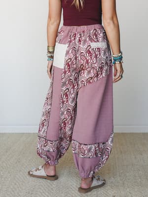 Shot from behind, these wide-leg harem joggers mix dusty mauve solid panels with a bold paisley patchwork and a lace-trim back pocket — styled with stacked bracelets and sandals for a well-traveled, free-spirited feel.