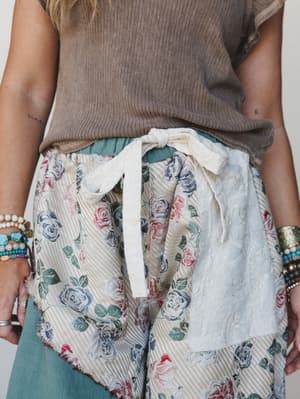 A close-up detail shot of mixed-fabric wide-leg pants with a floral tapestry front panel and drawstring waist, styled with a ribbed mocha tee and stacked boho bracelets — the kind of pants that feel like a story.
