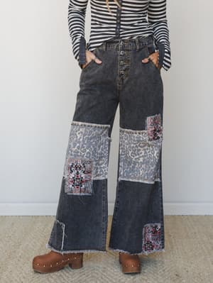 Wide-leg black denim jeans with mixed patchwork panels — leopard print, tribal/Aztec fabric, and raw fringe edges — styled with a striped long-sleeve top and chunky wooden clogs for a lived-in, expressive boho look.