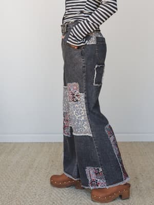 A side-profile shot of wide-leg black denim jeans covered in mixed patchwork — leopard print, Aztec-inspired fabric, and frayed edges throughout — styled with a striped long-sleeve and wooden clogs for a look that feels vintage-collected and unapolog