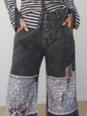 A close-up mid-shot of wide-leg black denim jeans with patchwork detailing — leopard print fabric panels meet frayed raw edges and studded trim at the knee, layered over a striped long-sleeve top for an effortlessly eclectic, vintage-boho feel.