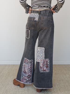 Back view of wide-leg dark wash denim pants with mixed patchwork panels — leopard print and boho tapestry — shot against a soft neutral studio wall with a striped crop top and brown clogs.