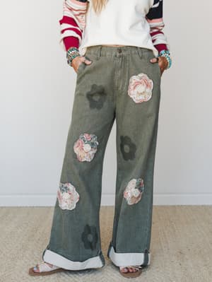 Wide-leg olive denim pants steal the show with mixed floral appliqués and tonal flower cutouts scattered across the legs, styled with a striped colorblock sweatshirt, stacked beaded bracelets, and rolled cuffs — the kind of pants that start conversat