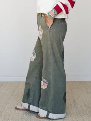 Wide-leg olive denim pants steal the show with floral appliqué patches and a rolled cuffed hem revealing a striped lining — styled with a cream and red striped sweater, turquoise bead bracelets, and sandals for an effortlessly expressive, vintage-boh