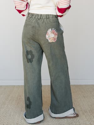 Shot from behind, a model wears wide-leg olive denim pants with tonal flower appliqués and a mixed floral patchwork detail at the hip, styled with a striped red-and-cream sweatshirt and rolled cuffs — the kind of pants that make a Saturday feel like