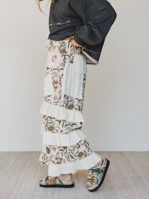 Person wearing floral pants with ruffles and a dark jacket on a plain background