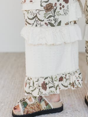 Person wearing floral pants with ruffled layers and matching floral sandals on a neutral background