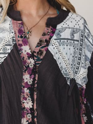 A close-up detail shot of a mixed-print patchwork top in charcoal, blush, and ivory — floral, paisley, and geometric panels stitched together with raw edges and ornate button closures, styled with a delicate gold charm necklace.