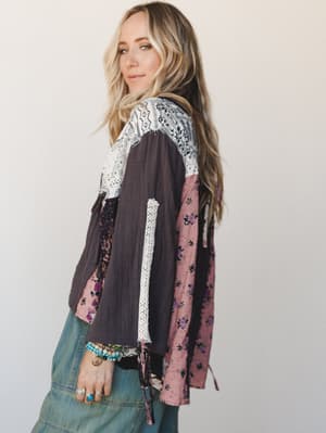 A side-profile shot of a model in a patchwork boho top — mixing charcoal gauze, dusty rose floral, and bandana-print panels with a lace trim detail — paired with light-wash wide-leg jeans and layered bracelets and rings that feel like she's been coll