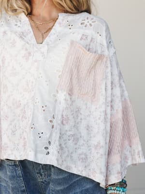 A close-up mid-shot of an airy, patchwork blouse in white and blush — layered with eyelet cutwork, toile print panels, and a crinkle lace pocket that feel like they were collected over time, styled with denim and turquoise bracelets for an effortless