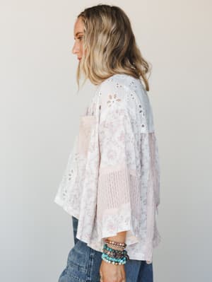 A side-profile shot of a flowy patchwork blouse in soft white and dusty rose, mixing eyelet, toile print, and textured panels — styled with denim and a stack of beaded bracelets for that effortlessly collected boho feel.