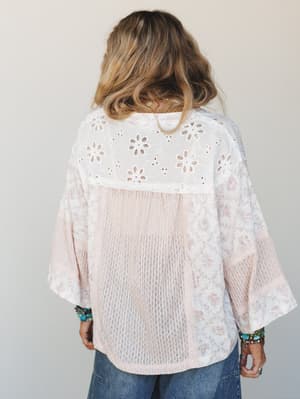 Shot from the back, this mixed-fabric blouse layers eyelet lace across the yoke over a soft blush floral print body with open-knit texture panels — styled with stacked turquoise bracelets and denim for that effortlessly collected, boho-everyday feel.