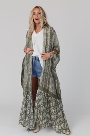 A woman with blonde hair wears the Paisley Tapestry Free Flow Duster Kimono in Olive over a white top and denim shorts, accessorized with layered necklaces and bracelets, standing against a plain white background.