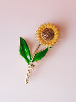 A sunflower hair clip laid flat against a soft blush background — gold-toned metal stem and leaves in painted enamel green, with amber rhinestone detail at the center of the bloom.
