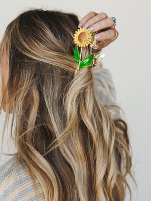 A close-up of wavy, sun-kissed hair held back with a gold and enamel sunflower claw clip, styled with stacked rings and soft nail art — the kind of detail that makes a whole outfit feel intentional.