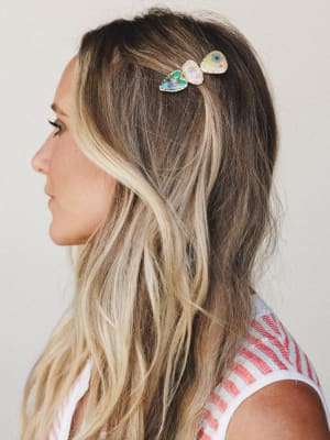 A side-profile close-up of a model wearing a multi-panel floral hair clip — three petal-shaped resin pieces in teal, blush, and soft yellow with gold trim — half-pinned into loose, beachy waves, paired with a glimpse of a red and white striped top.