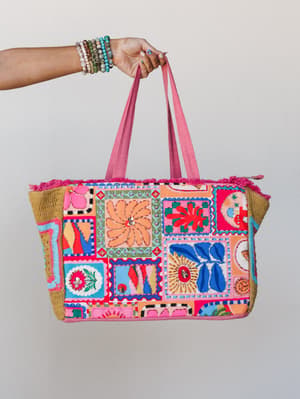 A hand holding up a large patchwork tote bag covered in bold embroidered and appliquéd panels — florals, suns, abstract shapes — with pink fringe trim, woven sides, and a stacked bracelet arm that looks like it belongs to the same person who'd pack t