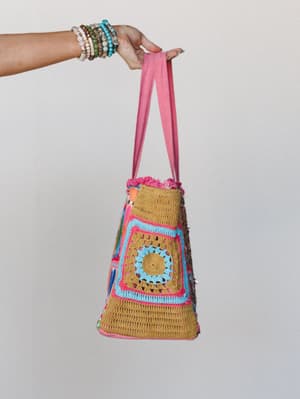 A stacked-bracelet wrist holds up a chunky crochet tote bag with a granny square panel, pink fringe trim, and a fabric strap — the kind of bag that makes your whole outfit feel more intentional.