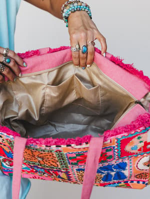 A close-up detail shot of a colorful embroidered tote being held open to show the interior, styled with stacked beaded bracelets, turquoise and silver rings — the kind of bag that has its own personality before you even pack it.