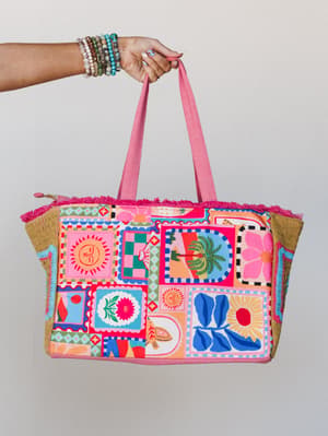 A colorful canvas tote bag held up by a stacked-bracelet wrist — the patchwork print is a full-on mood board of suns, palms, florals, and retro motifs with hot pink fringe trim and woven side panels.
