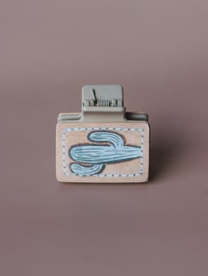 A square hair claw clip in muted sage, featuring a leather-look patch with a stitched cactus embroidery motif, shot clean against a dusty mauve background — the kind of little detail that makes a messy bun feel intentional.